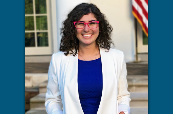 Hero graphic of Demitra Tomasides intern spring semester 2021 Smiling. She is wearing a white blazer and a bright dark blue shirt.