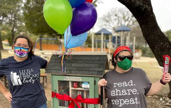 Next Chapter Book Club members stand next to a tiny library. Balloons are tied to the library. One member wears a shirt that says "Read. Share. Repeat"