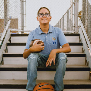 Kason Erwin sits on stairs and smiles. He is holding a football. A basketball is at his feet