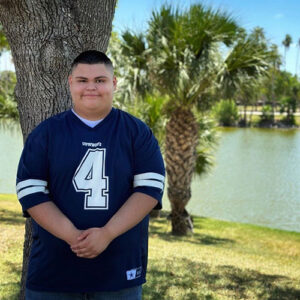 Matthew Leos stands in front of a tree and water, smiling. He is wearing a Dallas Cowboys jersey.