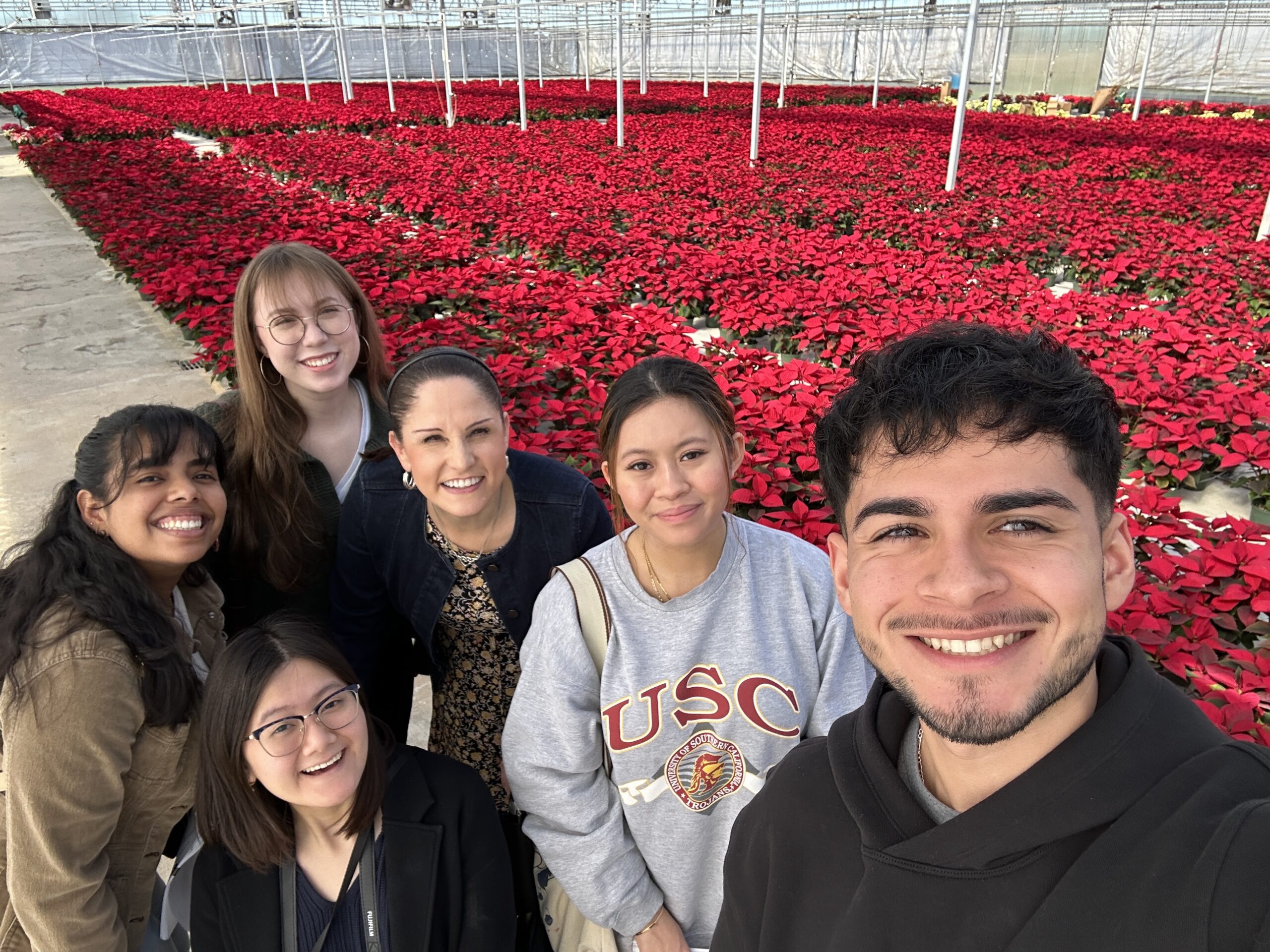 Group photo of Rice University grant project leaders and students in a greenhouse at the Brookwood Community in Brookshire, Texas. The photo includes Dr. Luz Garcini, Kathryn Gonzalez, Ling DeBellis, Sabrina Cuauro Cuauro, Beni Nguyen, and Ricardo Robles. Behind the group is an assortment of poinsettias that were grown by members of the Brookwood Community.
