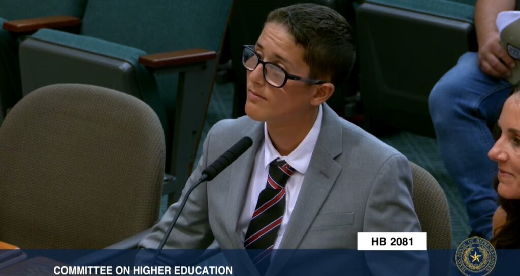 Kason Erwin testifying to the House Committee on Higher Education. Kason is sitting behind a desk, He has short dark hair and glasses and wears a suit and tie.