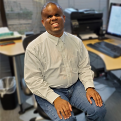 Jason Jones sits at an office desk in a chair. He smiles and wears a button up shirt.