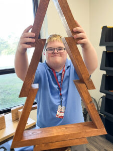 A smiling young man wearing glasses holding up a large, wooden Christmas tree decoration