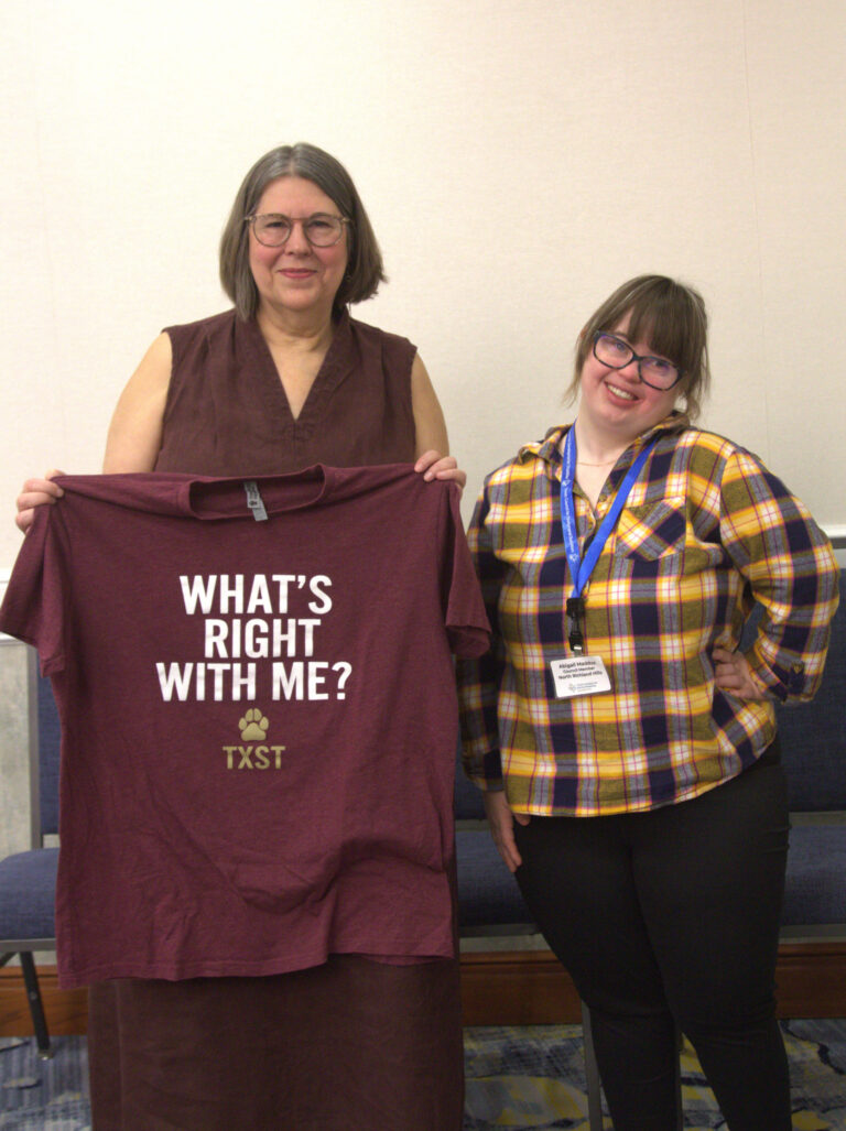 Two women standing together; both are smiling and both wear glasses. One holds up a T-shirt that says "What's Right With Me?". The other wears a yellow and dark grey plaid shirt and a name badge lanyard.