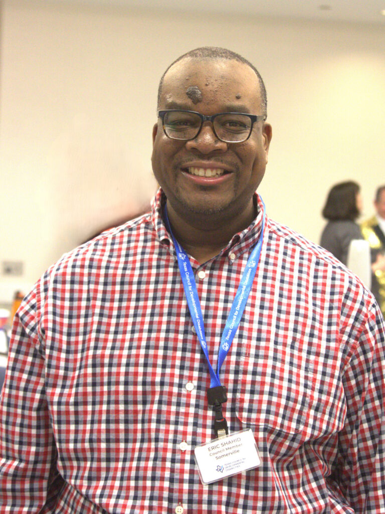 A smiling man wearing glasses, a name badge lanyard, and a red, black, and white plaid button-down shirt.