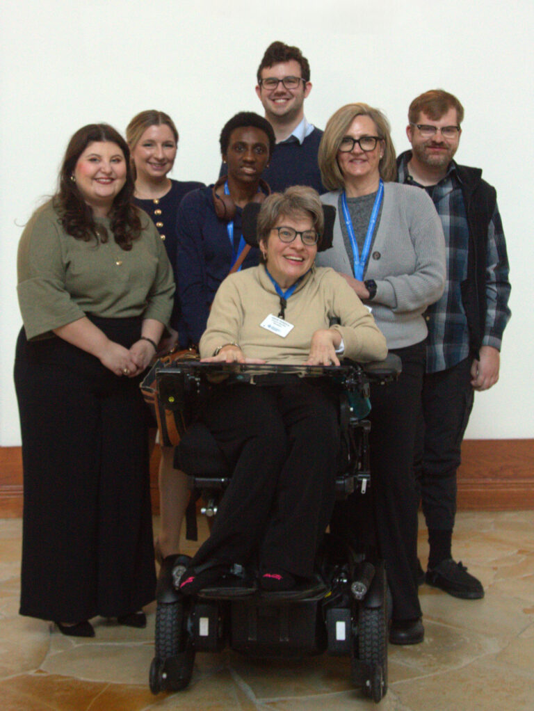 A group of Council members and Council meeting participants standing together. The group is arranged in rows against a light-colored wall. One person is in a power wheelchair in the center foreground.