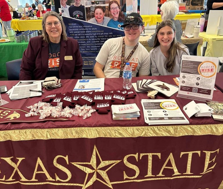 Three people — Dr. Tamara Shetron, Grant Zollinger, and Gloria Gilbert — sitting behind a table at an event inside a large indoor venue. All three are facing forward and smiling. The table is covered by a maroon tablecloth and holds stacks of brochures, flyers, buttons, and pens. Other booths, banners, and attendees are visible in the background.